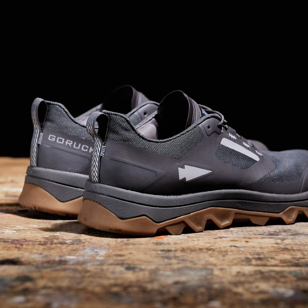 The "GORUCK" logo is featured on gray Mackall - Forged Iron + Chiseled Stone + Gum shoes with tan soles, displayed on a wooden surface. Designed for all-terrain rucking, their triple compound outsole ensures stability and grip for any adventure.
