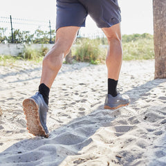 While jogging on sandy terrain near a palm tree, a person wears black socks, gray shorts, and GORUCK's Mackall in Forged Iron + Chiseled Stone + Gum, which have an all-terrain triple compound outsole.