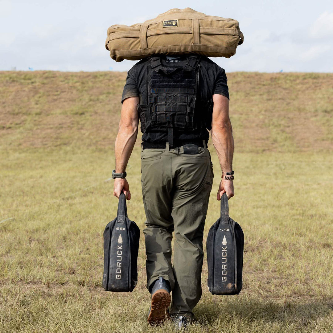 Wearing GORUCK's Mackall - Forged Iron + Chiseled Stone + Gum shoes, a person confidently strides across the grass with a large bag on their shoulders and two weights in hand, heading for a hill.