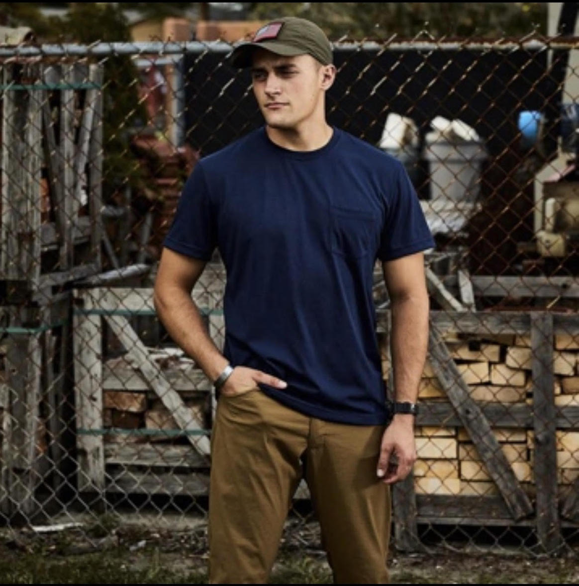 Man in navy t-shirt and khaki pants stands by a chain-link fence with stacked wood and crates behind him.