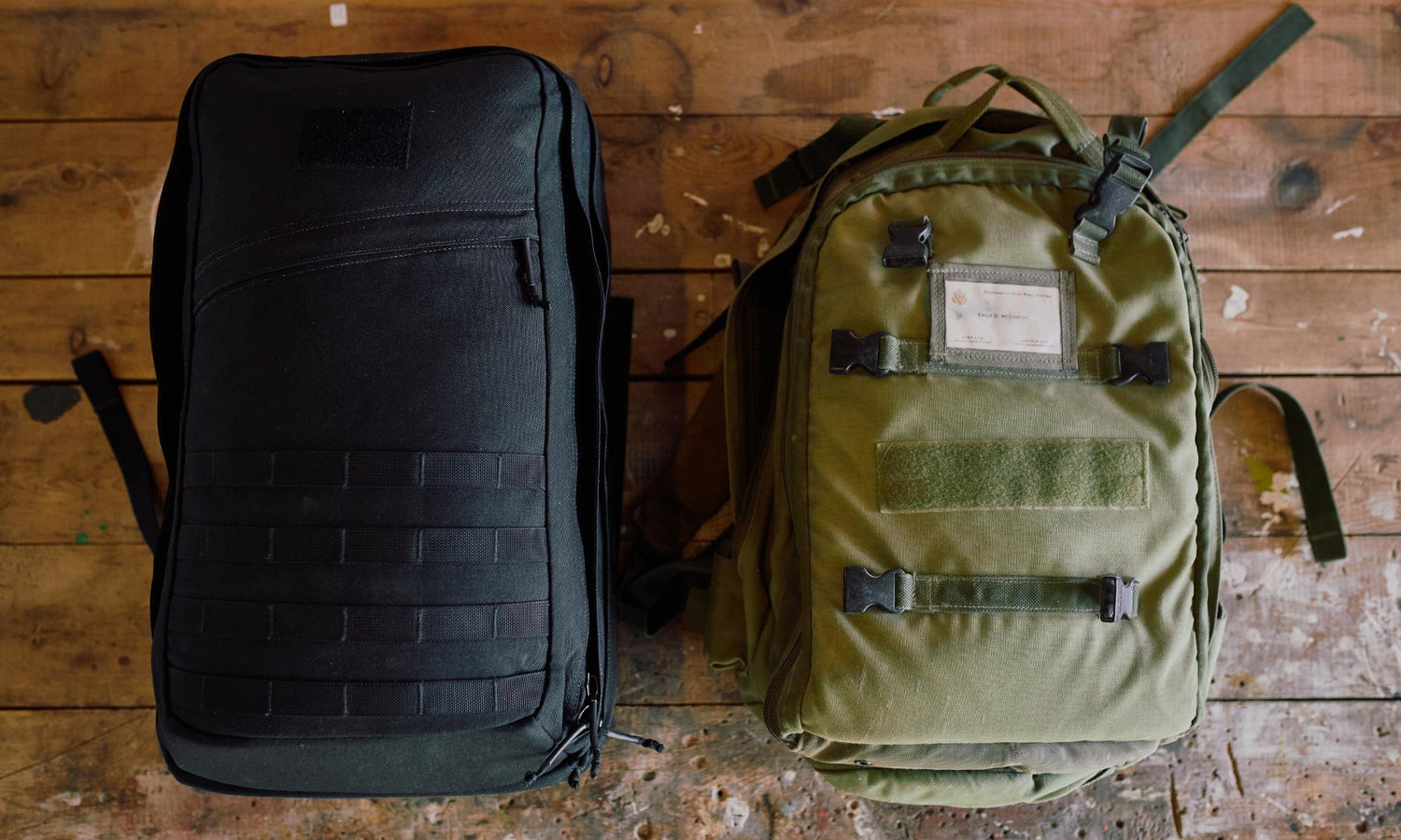 A black backpack and a green backpack side by side on a worn wooden surface.