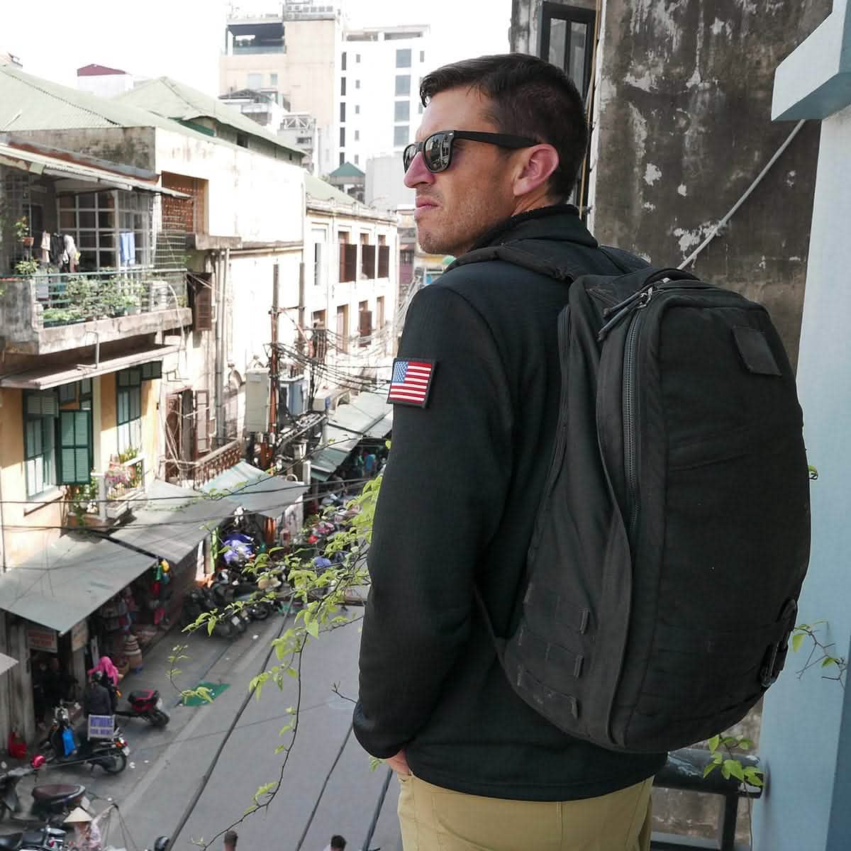 A person with a sleek GR1 USA rucksack and sunglasses from GORUCK surveys the bustling urban street from a balcony.