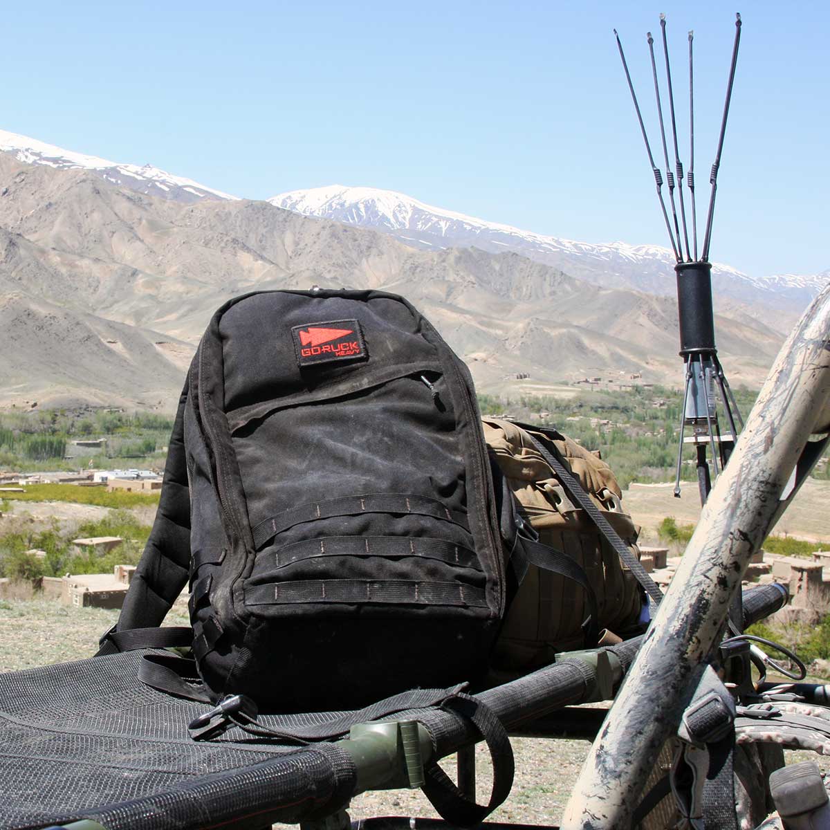 Black backpack on military gear with mountains and green valley in the background under a clear blue sky.