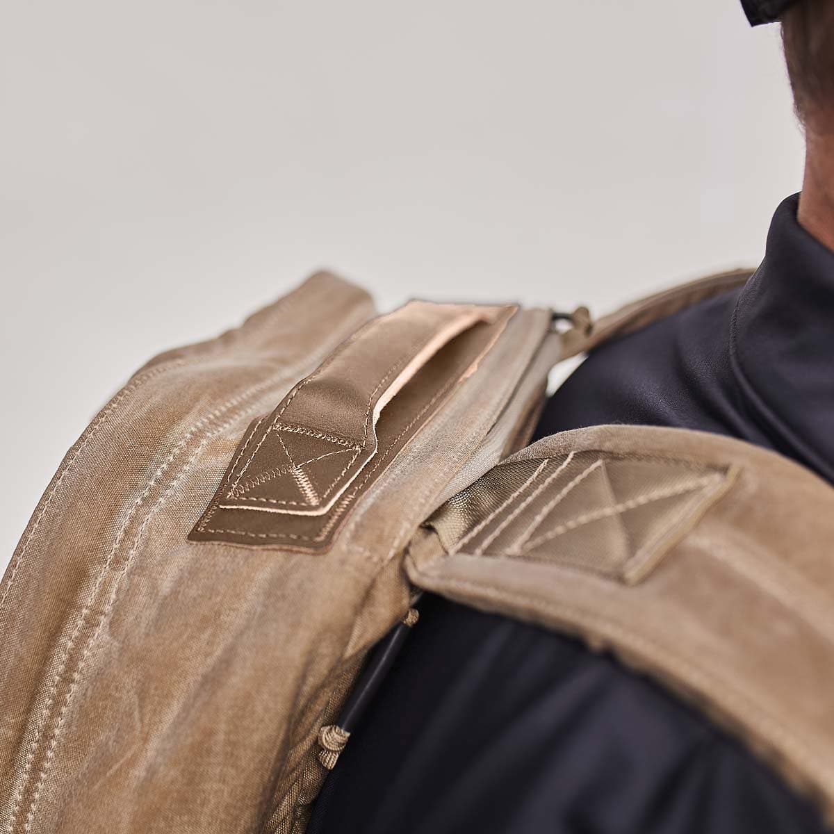 Close-up of a person wearing a tan backpack with reinforced stitching on the straps.