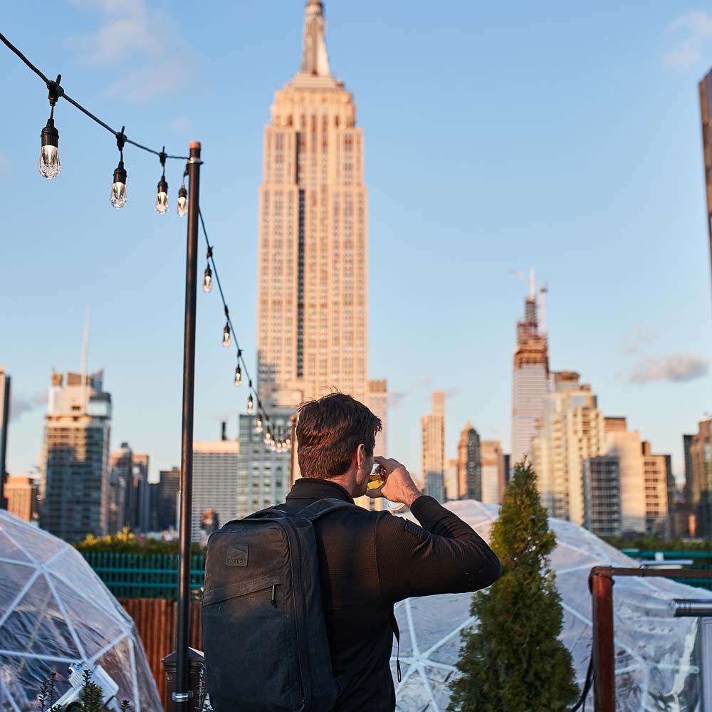 A person with a backpack drinks on a rooftop with the Empire State Building in the background at sunset.