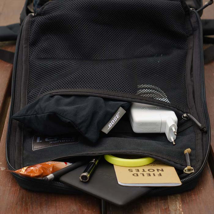 Open backpack with mesh pocket, charger, Bose case, notebook, pen, and other small items inside.