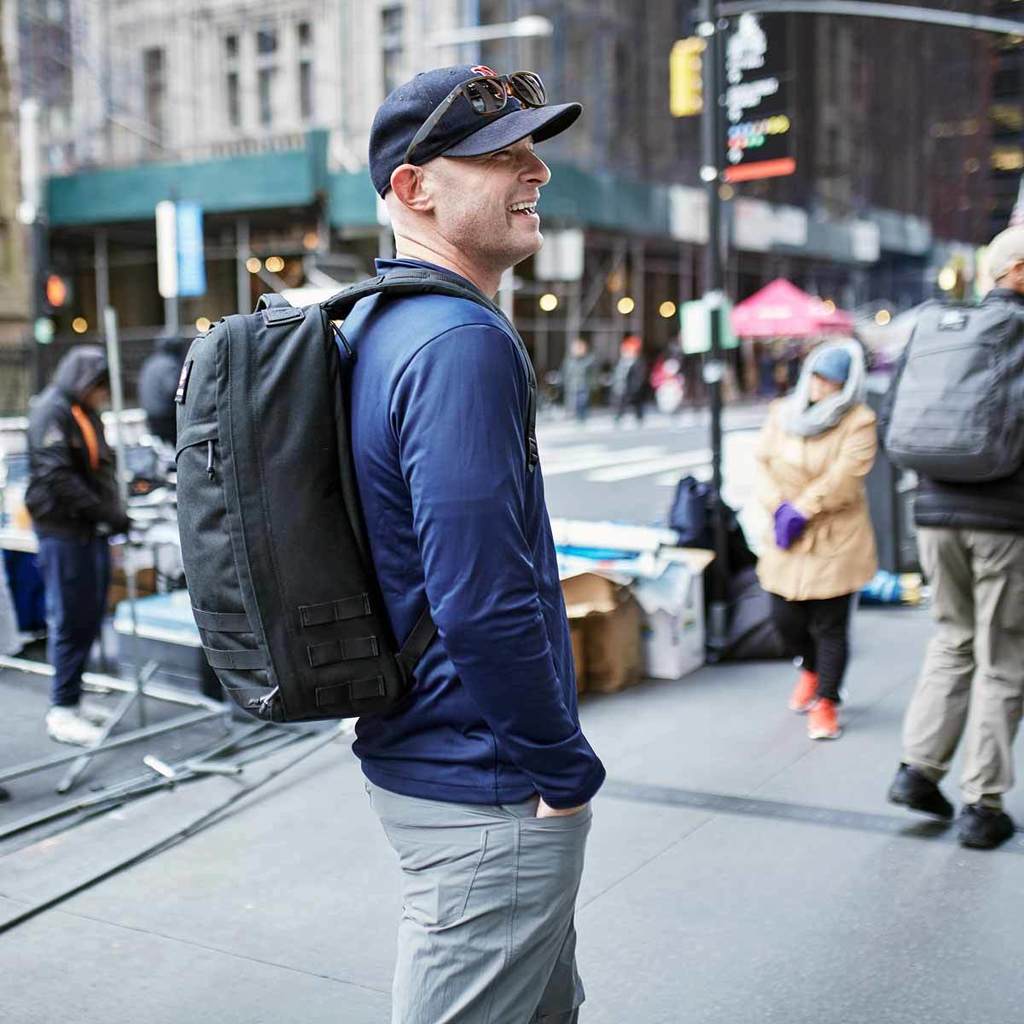 A smiling man in a blue jacket and cap stands on a city street, carrying a large black backpack.