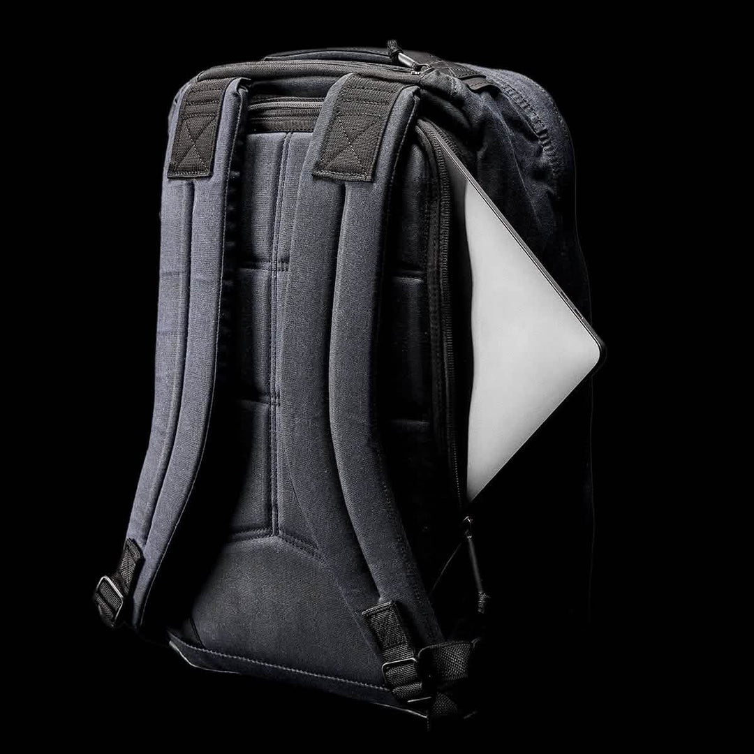 The GR1 USA - Heritage by GORUCK is a black waxed canvas rucksack with padded straps and a partially open side zipper pocket revealing a laptop, set against a black background, highlighting its modern design and heritage-inspired durability.