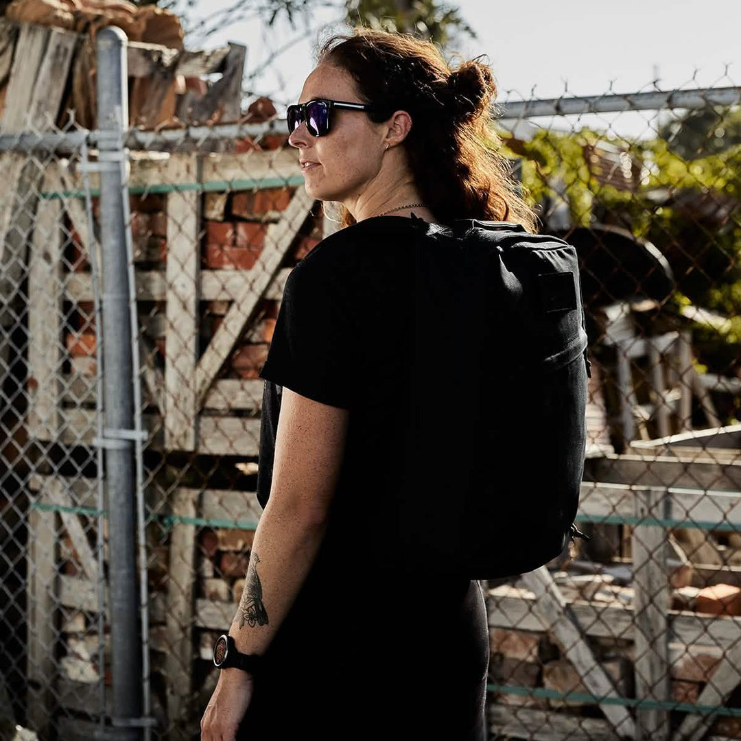 A person wearing sunglasses and a GORUCK GR1 USA - Heritage rucksack is standing by a chain-link fence, with piled bricks forming an industrial backdrop.