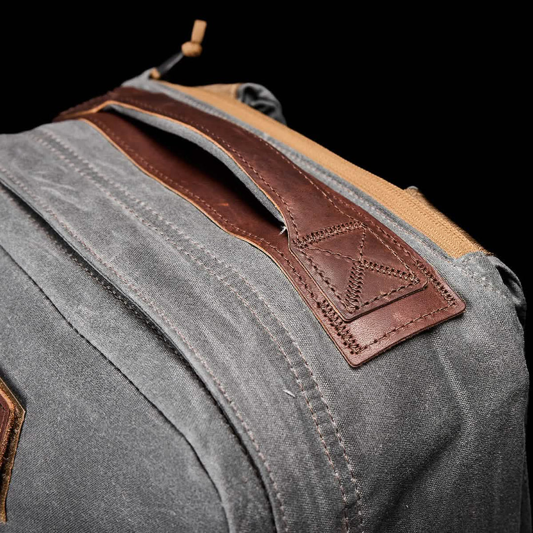 Close-up of a GR1 USA - Heritage Rucksack in gray fabric with a brown leather handle and tan accents, crafted from durable waxed duck canvas by GORUCK, set against a black background.