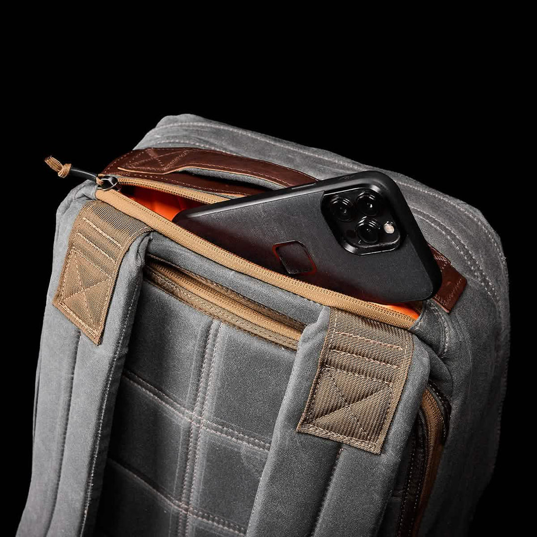 The GORUCK GR1 USA - Heritage Backpack, made from waxed duck canvas, features brown zippers and straps. It sits partially open with a black smartphone tucked in its pocket.