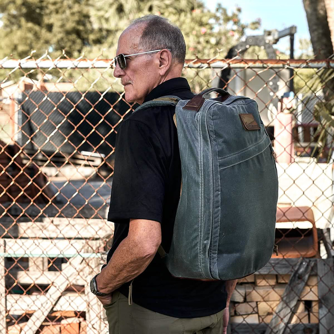 A man wearing sunglasses and a GORUCK GR1 USA - Heritage backpack stands by a wire fence, with trees and construction materials in the background.