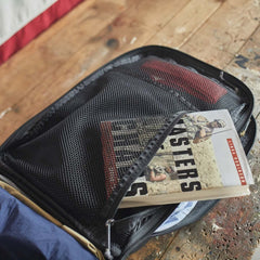 An open GR2 rucksack by GORUCK rests on a wooden table, showcasing a mesh pocket holding a book titled *Masters of Blinds*. Its compact design is carry-on compliant and comes with the assurance of a Scars Lifetime Guarantee, making it an ideal travel companion.
