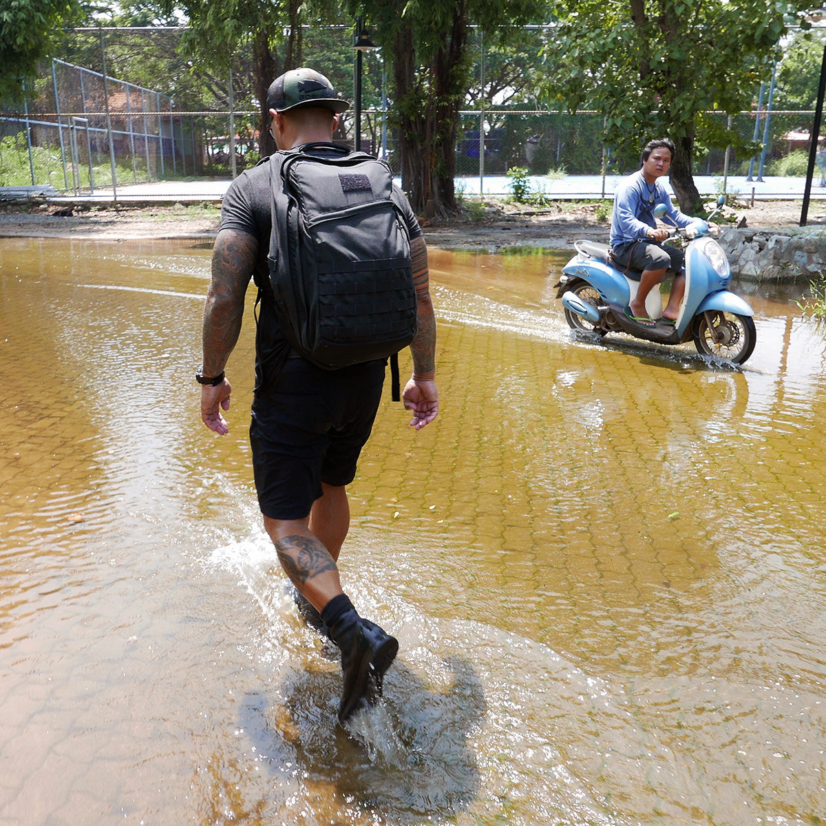 Man with a backpack walks through floodwater as a person on a scooter rides nearby.