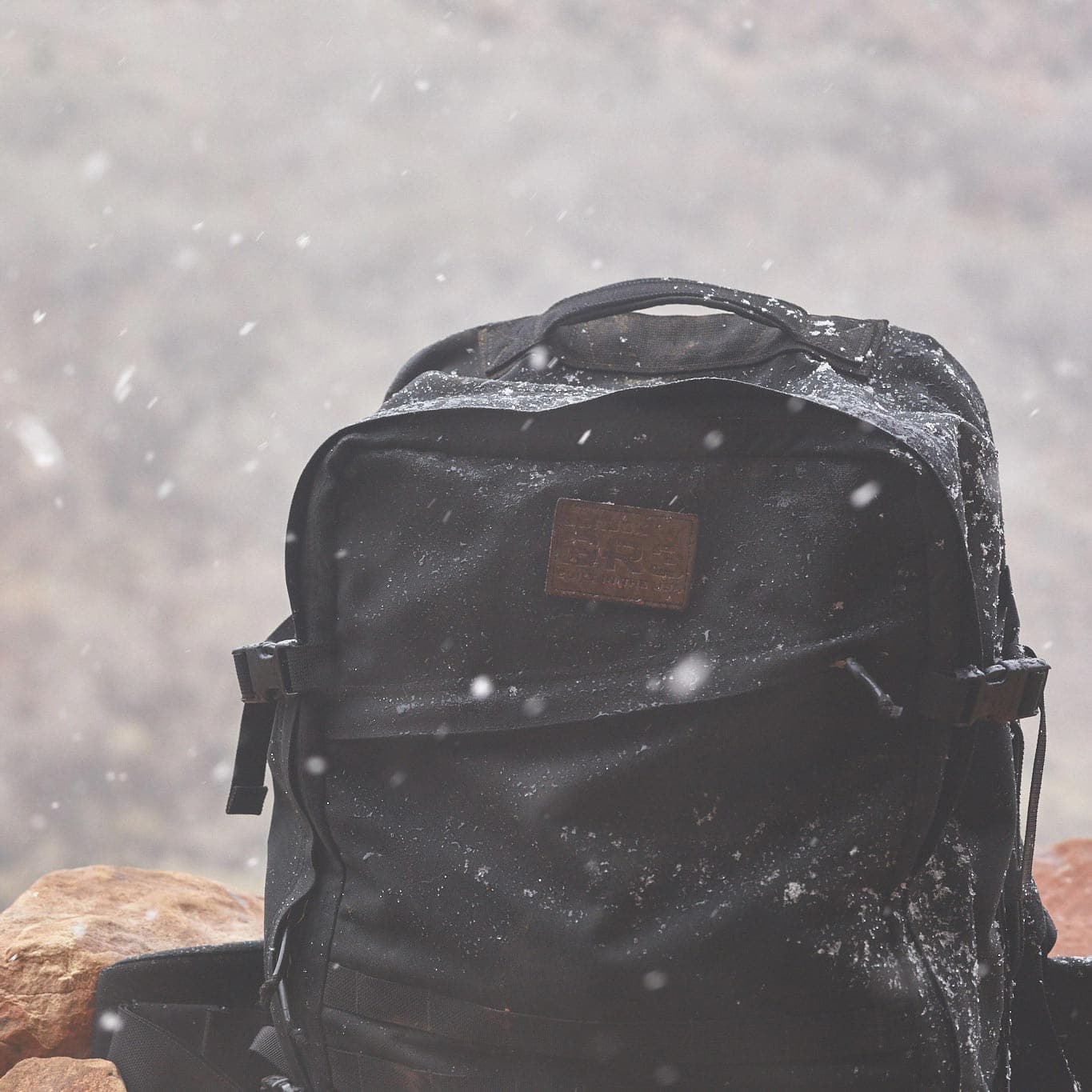 A black backpack sits on rocks, dusted with snow, as snowflakes fall around it.