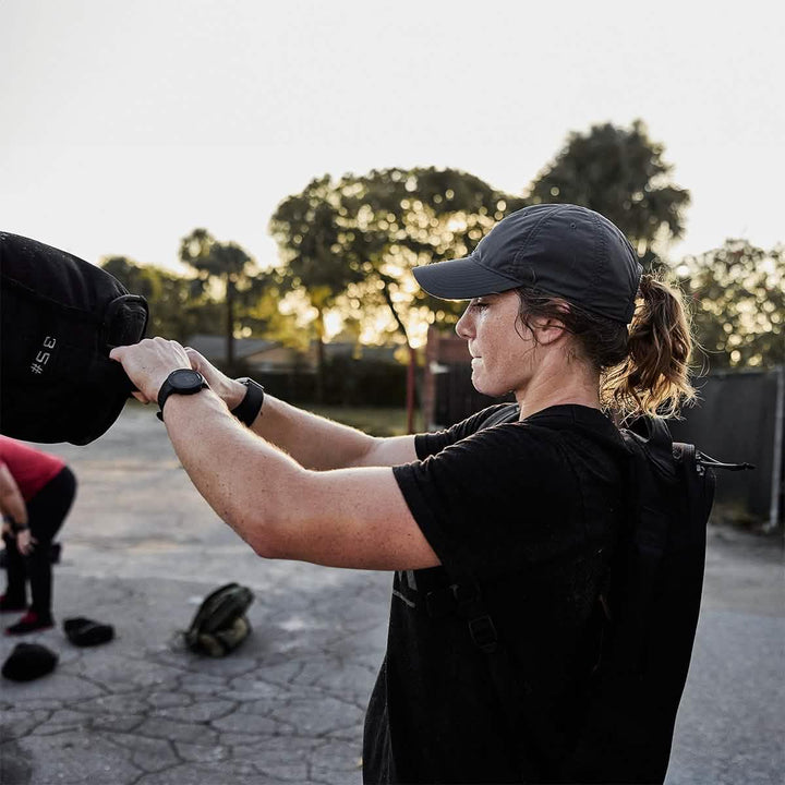 Woman lifting GORUCK sandbag outdoors, wearing backpack and cap during rucking workout