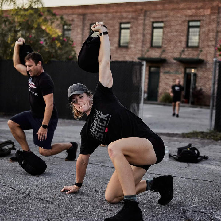 Two people training outdoors with GORUCK rucksacks, performing fitness exercises on pavement.