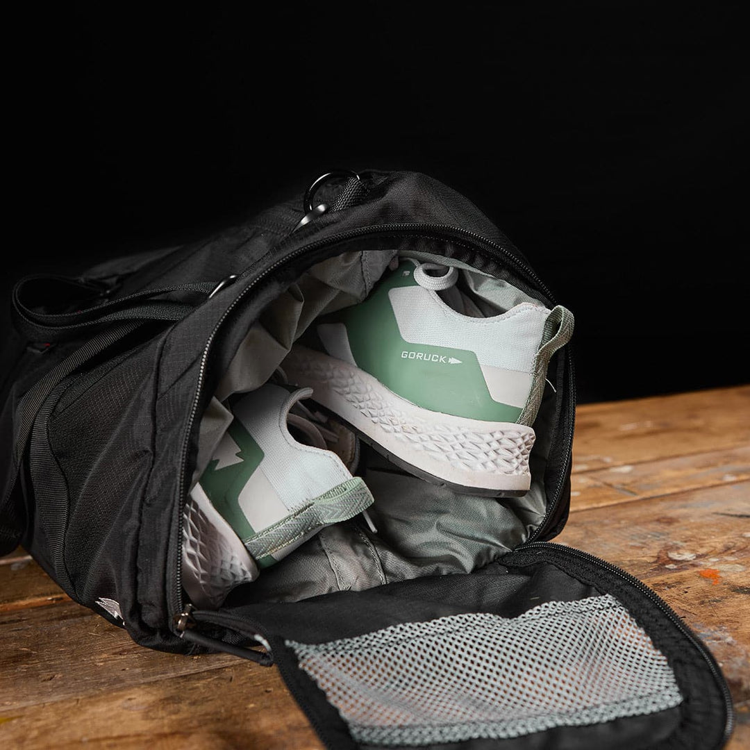 A GORUCK Gym Bag - Ripstop ROBIC® in black opens to reveal green and white sneakers neatly tucked inside, resting on a wooden surface.