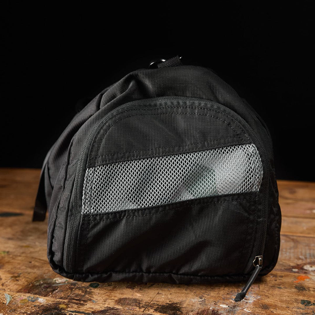 The Gym Bag - Ripstop ROBIC® by GORUCK, designed in a black hue and crafted from durable ripstop nylon, showcases a mesh pocket as it elegantly rests on a wooden surface against a dark background.
