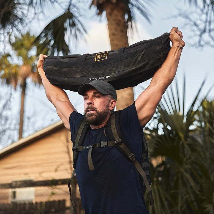 Man outdoors lifting GORUCK sandbag overhead, wearing rucking gear and GORUCK cap.