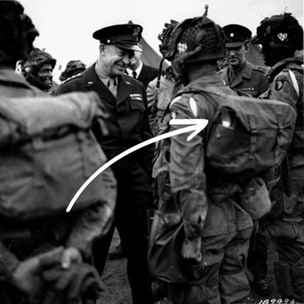 Military officer smiling and speaking to paratrooper; white arrow points to the soldier's uniform patch.