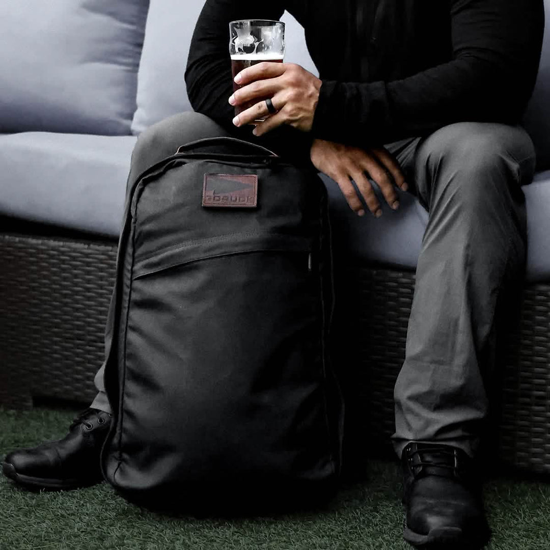 A person sits on a sofa holding a drink, with the GORUCK GR1 USA - Heritage, a renowned and durable waxed canvas rucksack, on the floor in front.