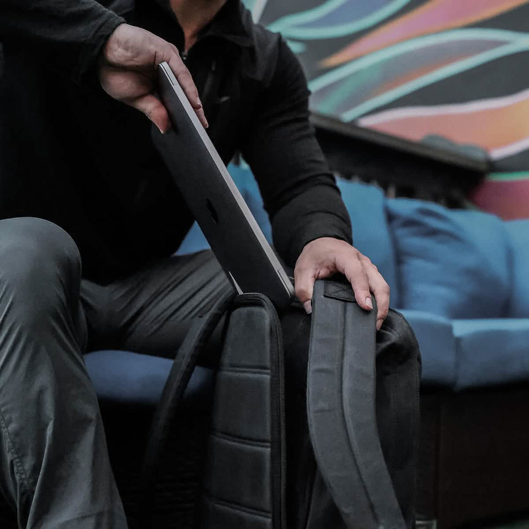 A person places a laptop into the GR1 USA - Heritage by GORUCK, resting on a couch with blue cushions and colorful wall art, ensuring both style and durability.