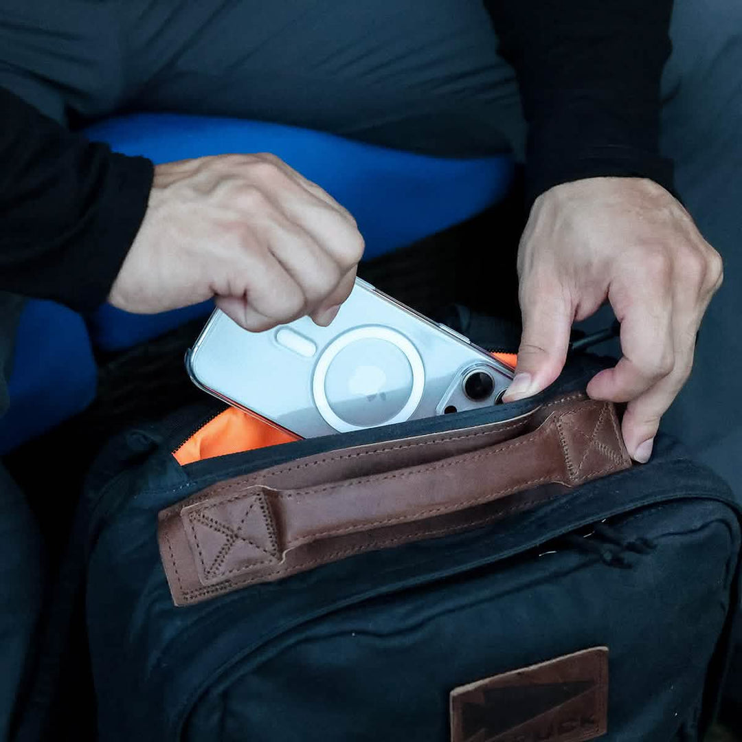 Someone gently stores a smartphone in a GORUCK GR1 USA - Heritage rucksack made from durable waxed canvas, with an orange lining and rich brown leather accents.