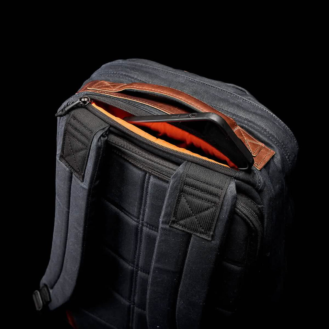 The GR1 USA - Heritage by GORUCK is a black rucksack with an open top zipper revealing a striking orange interior and visible tablet. Made from durable waxed canvas, it combines style and functionality for modern adventurers.