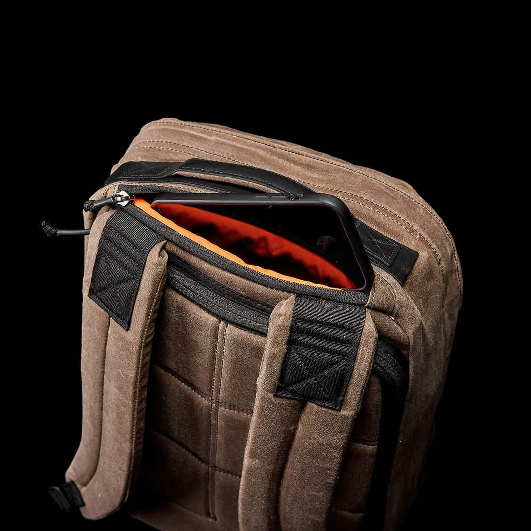 The GR1 USA - Heritage by GORUCK showcases a retro canvas rucksack with a brown exterior, complete with a phone peeking out from its padded pocket, all set against an elegant black background.