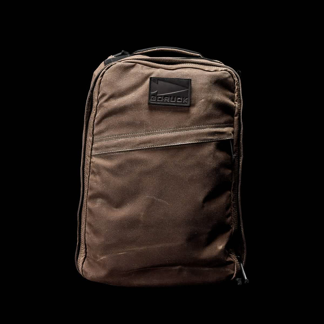 The GR1 USA - Heritage by GORUCK is a brown waxed duck canvas backpack with a front zipper pocket, set against a black background.