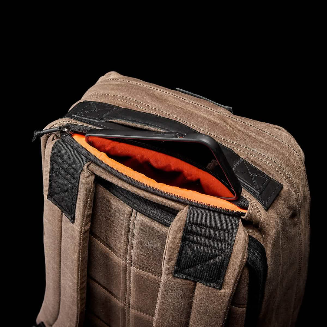 The GR1 USA - Heritage by GORUCK is a retro canvas rucksack made from waxed duck canvas, showcasing a brown body with an orange-lined pocket partially open against a black background.