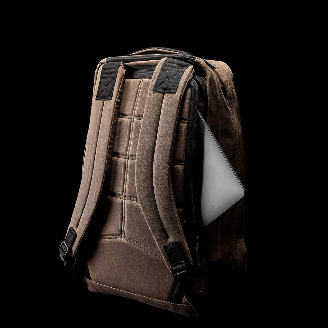 The GORUCK GR1 USA - Heritage is a retro-style rucksack featuring a brown design with padded straps, a laptop partially visible in an external side pocket, and crafted from durable waxed duck canvas for a timeless look. All set against a black background.