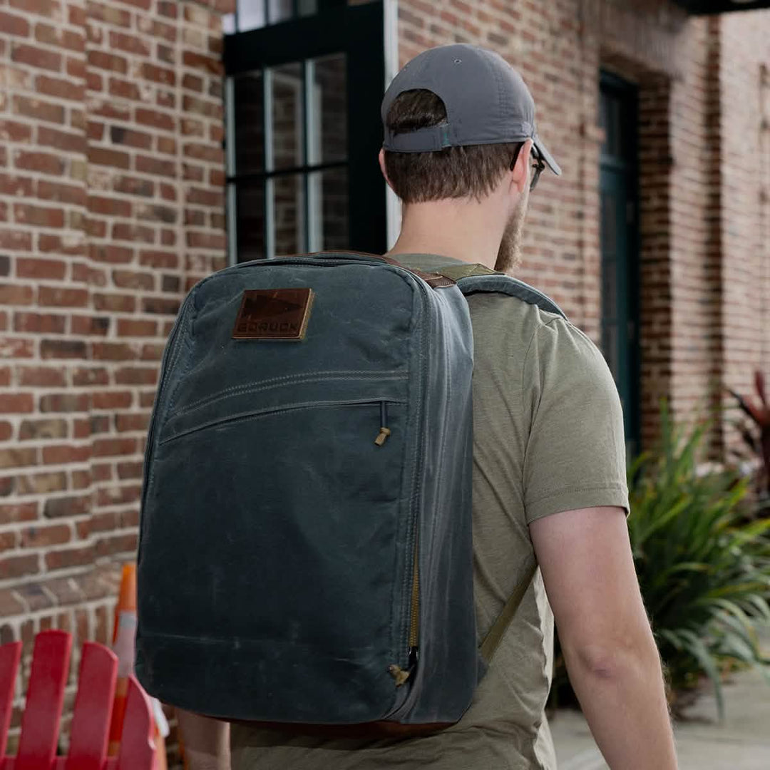 A man with a gray backpack and cap strolls outside by a brick building, his GR1 USA - Heritage from GORUCK subtly contrasting against the rustic backdrop.