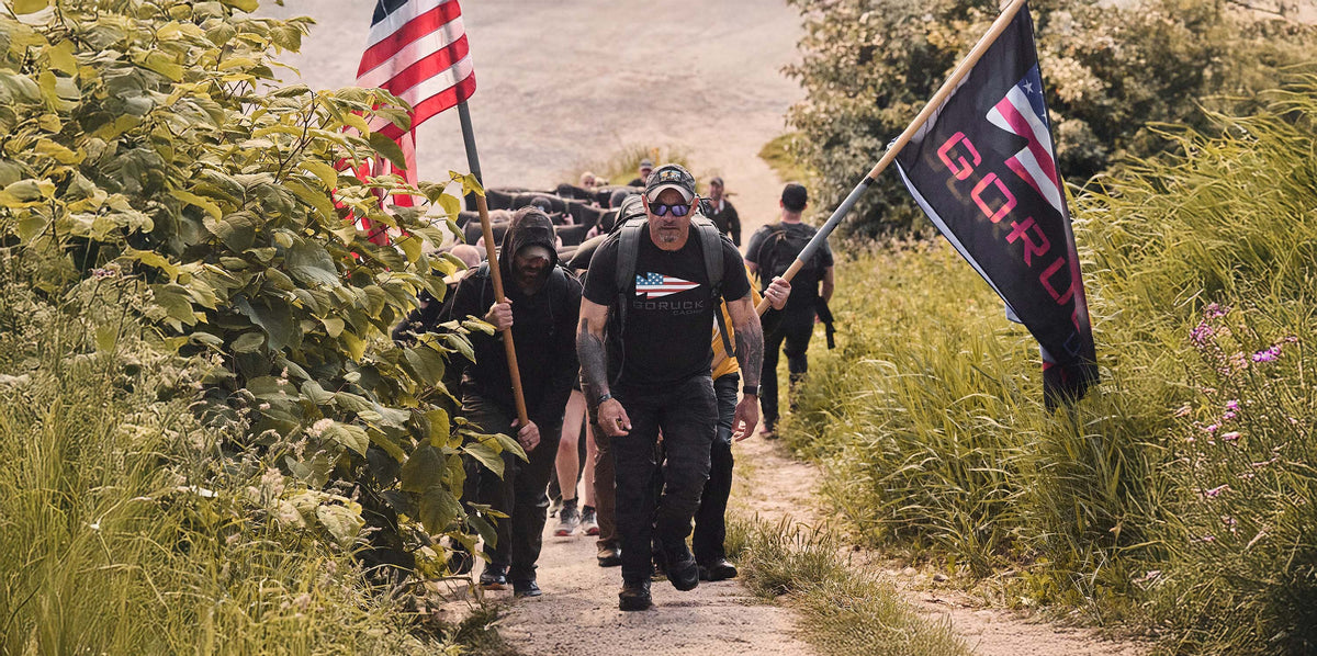A group of people hike up a path, carrying American and GORUCK flags, surrounded by greenery.