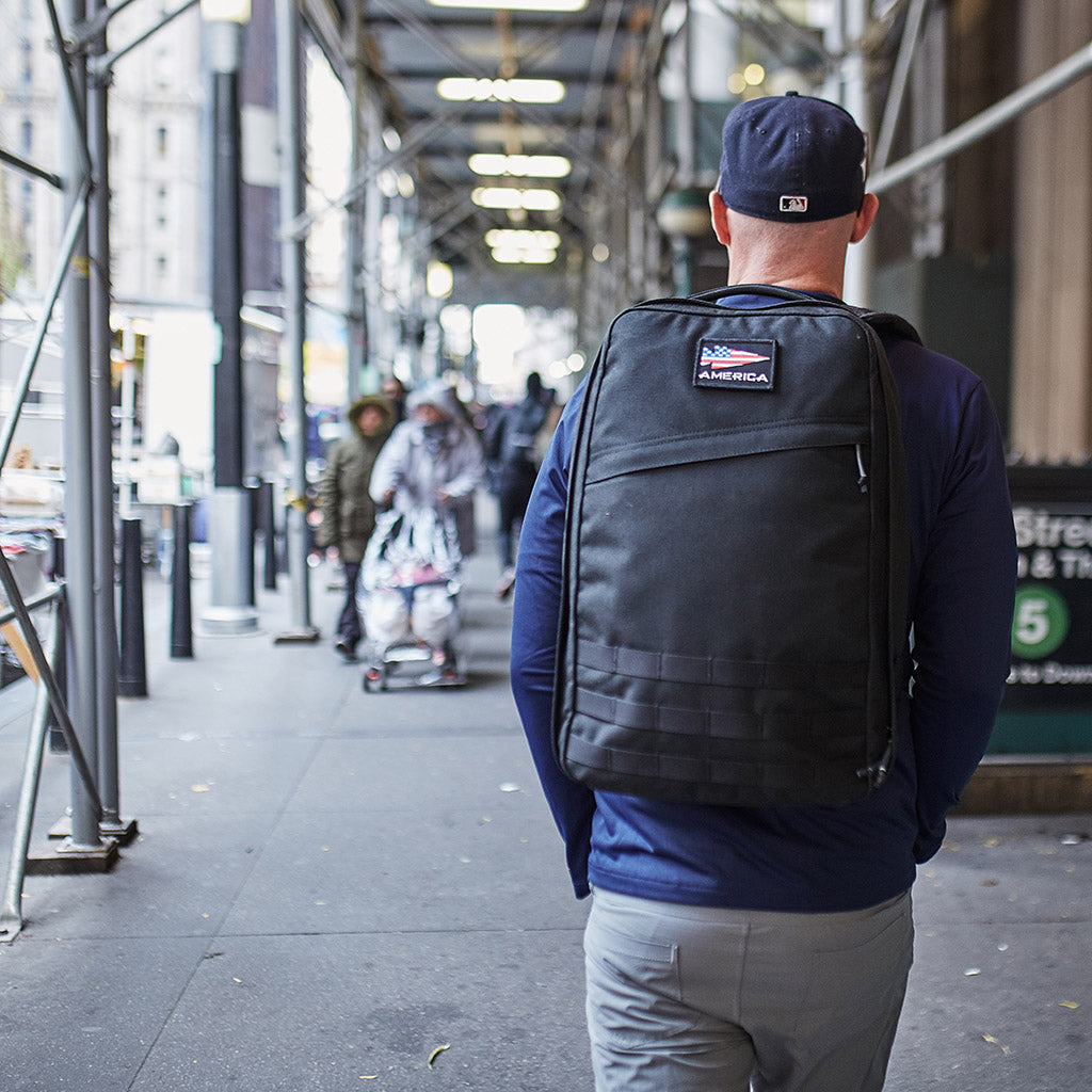 A man wearing a black backpack walks down a city sidewalk near a subway entrance.