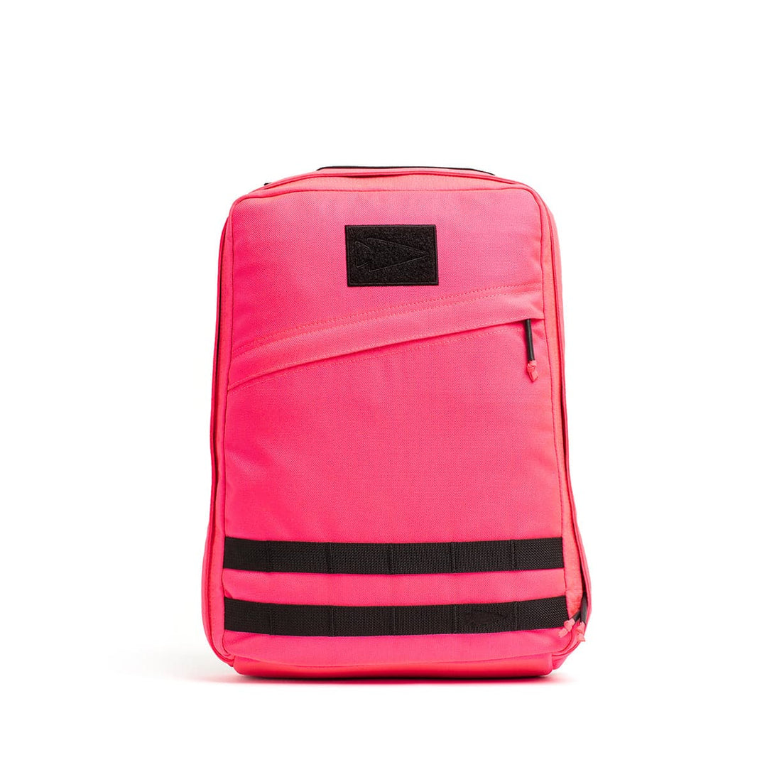 The GR0 - Cordura backpack by GORUCK showcases a vibrant pink color and a minimalist EDC design. It features a black rectangular patch, a vertical zip pocket, and two horizontal black straps near the bottom. A plain white backdrop emphasizes its sleek structure.