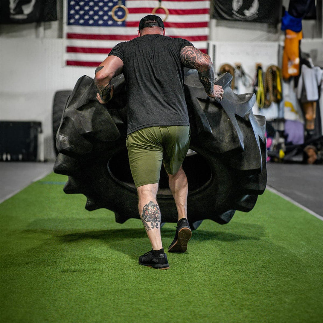 A tattooed man flips a large tire in a gym, showcasing his GORUCK Men's Ballistic Trainers - HULK. He is dressed in a black shirt and green shorts, sporting the latest in functional fitness footwear. An American flag and gym equipment can be seen on the green turf surface in the background.
