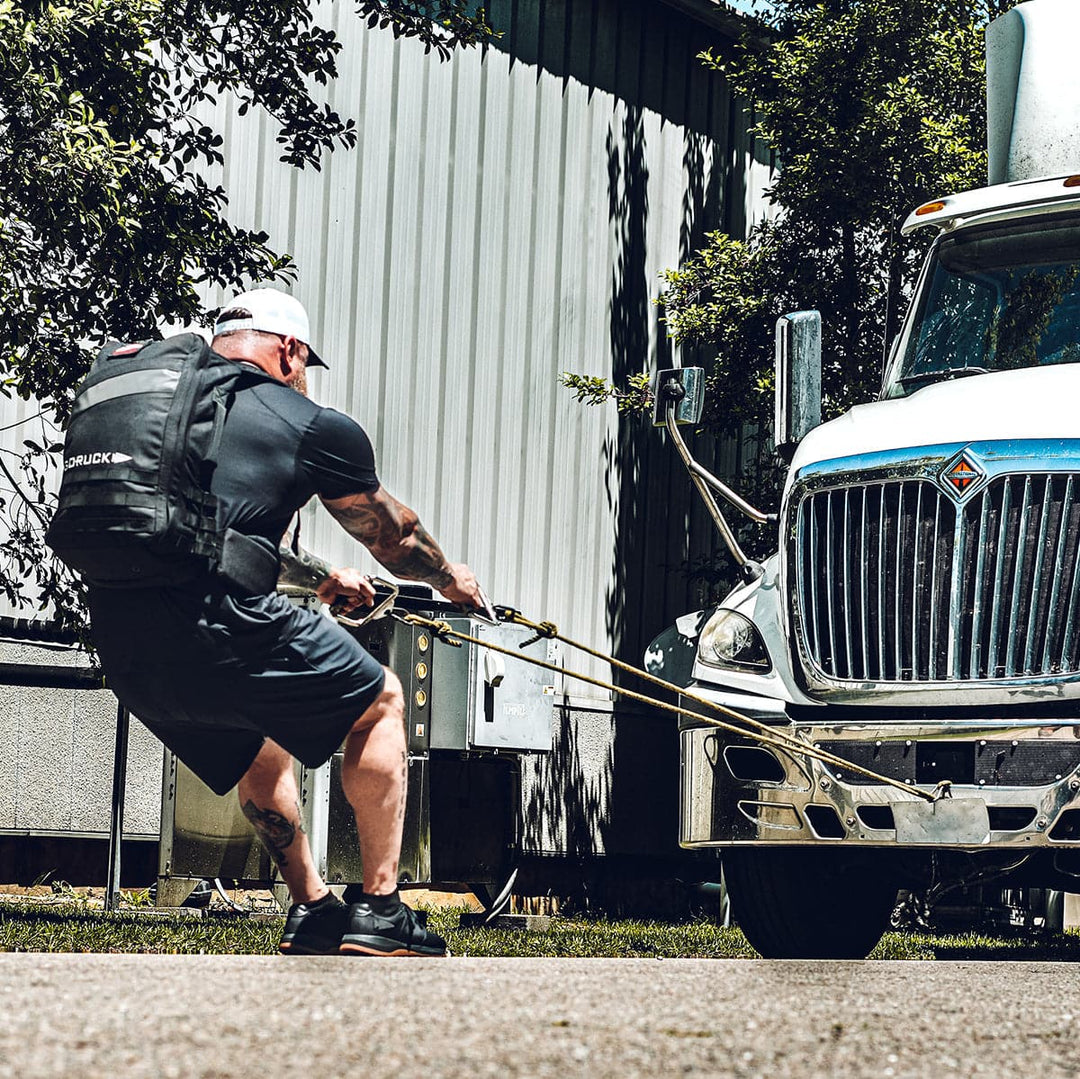 A muscular individual, outfitted in GORUCK's Men's Ballistic Trainers - HULK and athletic gear, pulls a large truck using ropes while wearing a weighted vest. The scene, highlighting the functional fitness qualities of this footwear, unfolds outdoors beside a metal building with trees in the background.