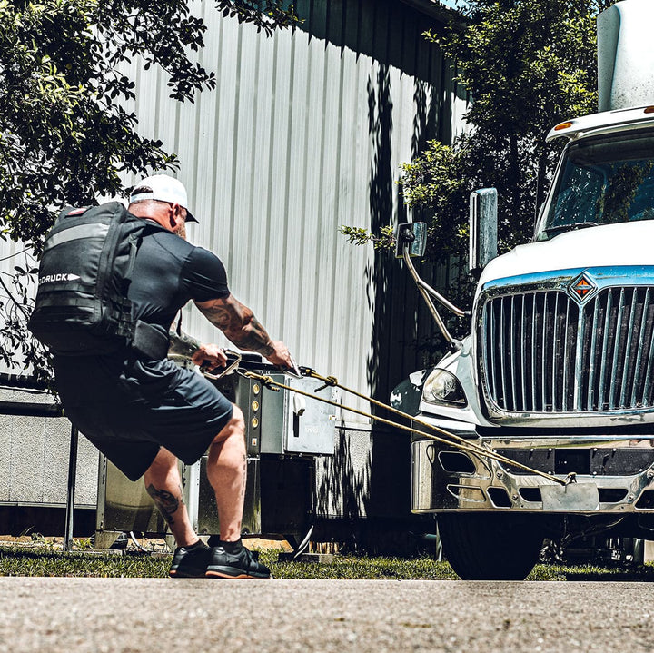 A muscular individual, outfitted in GORUCK's Men's Ballistic Trainers - HULK and athletic gear, pulls a large truck using ropes while wearing a weighted vest. The scene, highlighting the functional fitness qualities of this footwear, unfolds outdoors beside a metal building with trees in the background.