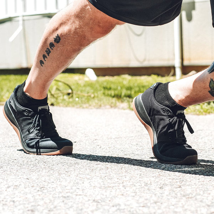A person wearing GORUCK Men's Ballistic Trainers - HULK and shorts jogs on a sunlit path. The tattooed leg is visible, and the scene suggests an active, outdoor atmosphere.