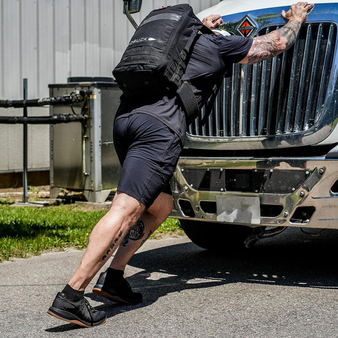 A man in black athletic gear and a backpack, wearing GORUCK's Men's Ballistic Trainers - HULK, pushes against the front of a large truck on a street. The truck's shiny grille and the man's tattoos are visible. A building and grass are in the background.