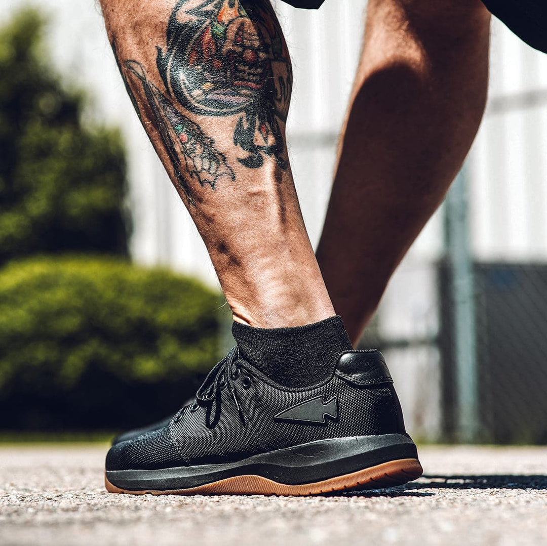 A person with tattooed legs steps forward wearing GORUCK's Men's Ballistic Trainers - HULK, featuring black design and brown soles. The scene is outdoors, with blurred greenery and a fence in the background, perfectly showcasing the blend of style and function in these workout shoes.