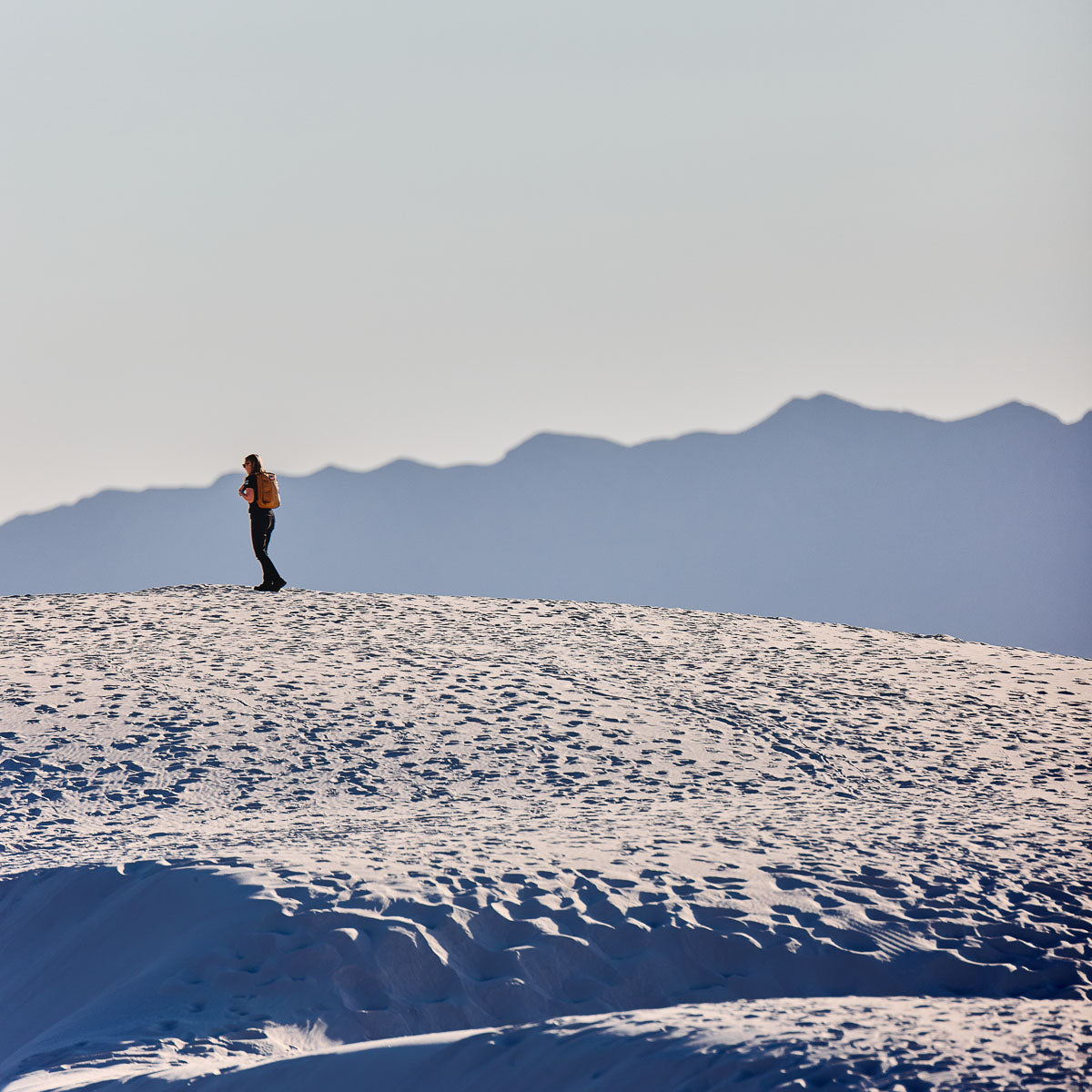 Person with a backpack stands on a sandy dune with mountains in the background under a clear sky.