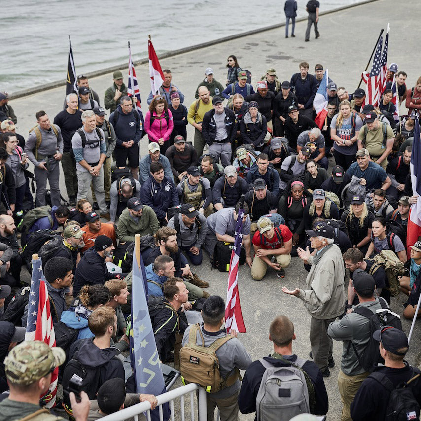 A large group of people with flags gather outdoors by a waterfront, listening to a man speaking.