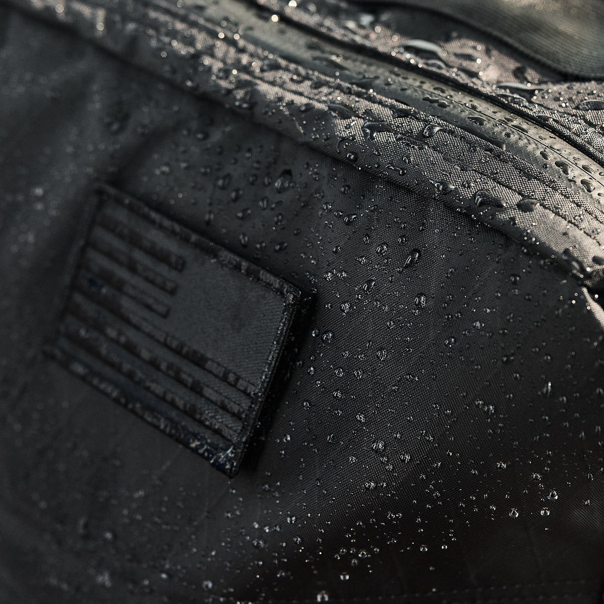 Close-up of water droplets on a black fabric bag with a stitched patch and a visible zipper.