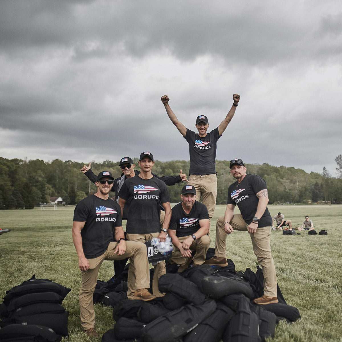 Six people pose outdoors on grass, some standing and some kneeling on sandbags, wearing GORUCK shirts and hats.