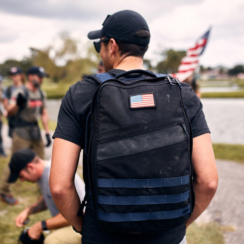 Man wearing a backpack with a U.S. flag patch, standing outdoors with others and an American flag in the background.