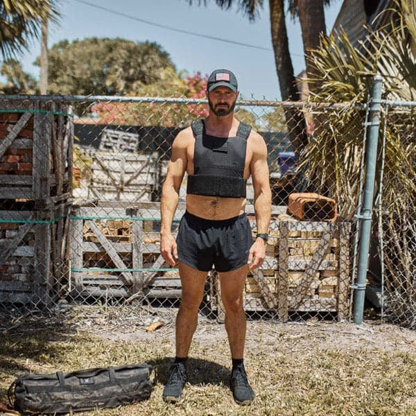 Man wearing a weighted vest and shorts stands outdoors near a fence, with workout gear on the ground.