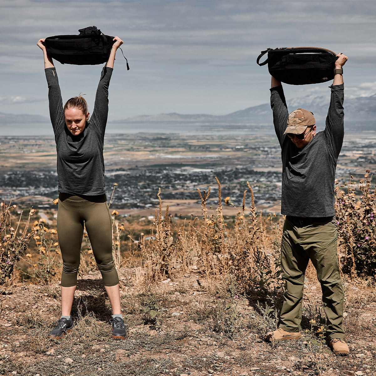 Two people outdoors holding backpacks overhead, standing on a dirt trail overlooking a scenic landscape.